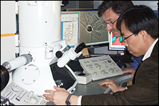 Specialists inspecting a high power microscope in the Nanoscale Imaging Spectroscopy & Properties Laboratory (AIMLab)