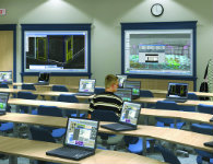 Lecture style classroom space with laptops set on tables in front of a projector. a single student sits at one of the tables.