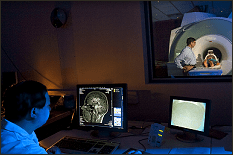 Looking over the shoulder of a technician in the control room of an MRI, looking through the window into the room with the machine itself
