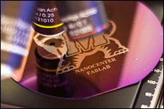 Close up of the specimen tray under a microscope looking at a wafer imprinted with an image of tested and the words: NanoCenter FabLab